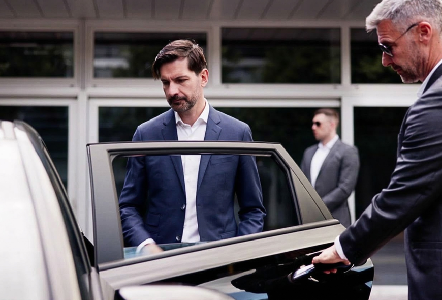 Close protection driver opening a car door while monitoring surroundings, highlighting the added security awareness and protective role compared with a standard chauffeur service.