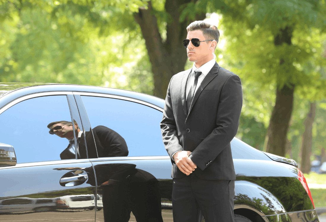 Close protection officer in a dark suit and sunglasses standing alert beside a luxury car, conveying professional personal security, discretion, and executive protection for high-net-worth individuals and family offices.